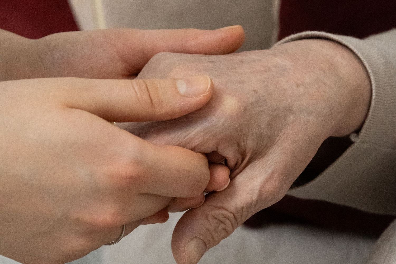 A younger hand holds an older woman's hand where it rests on the arm of a chair