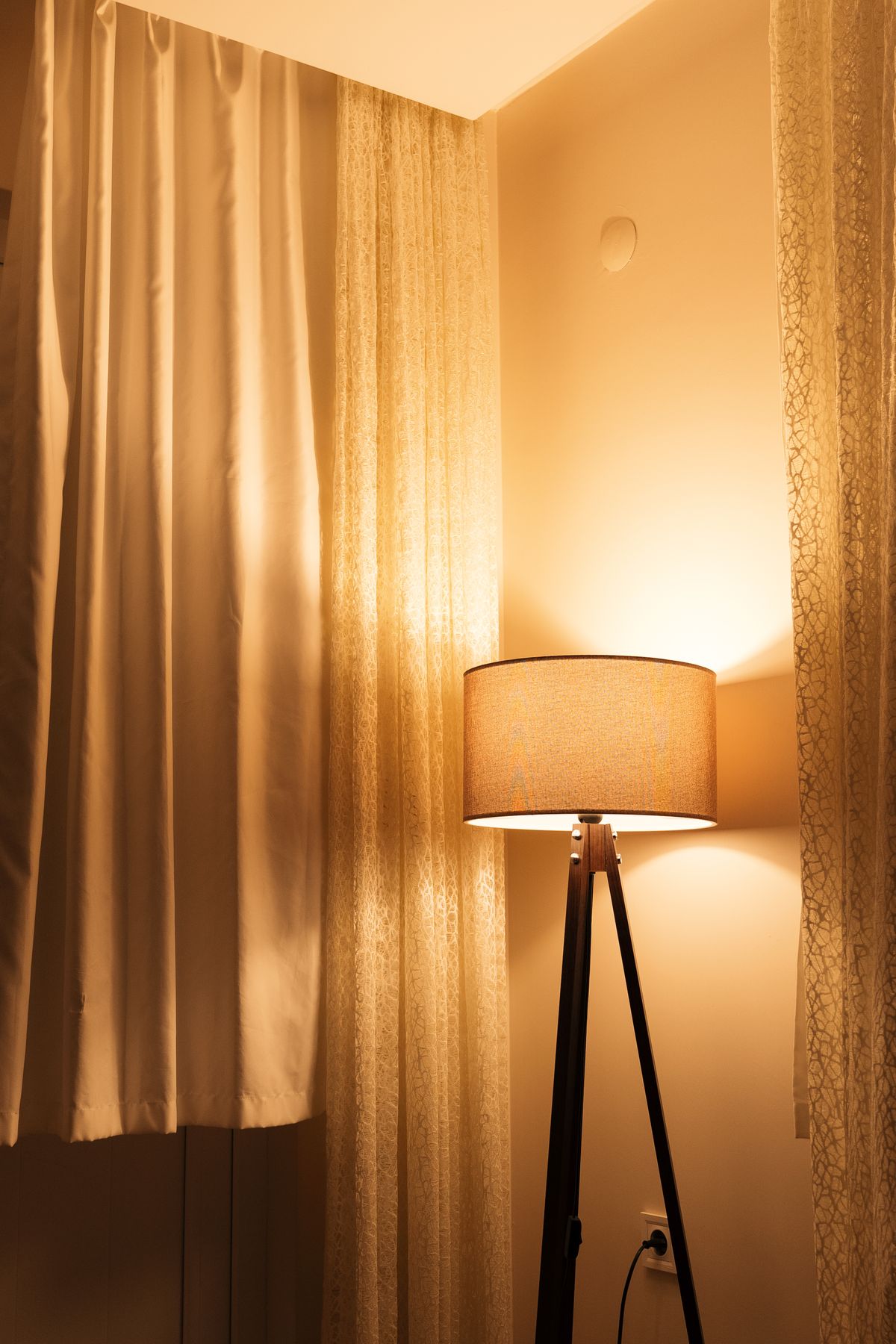 A floor lamp glows in the corner of a curtained room at night